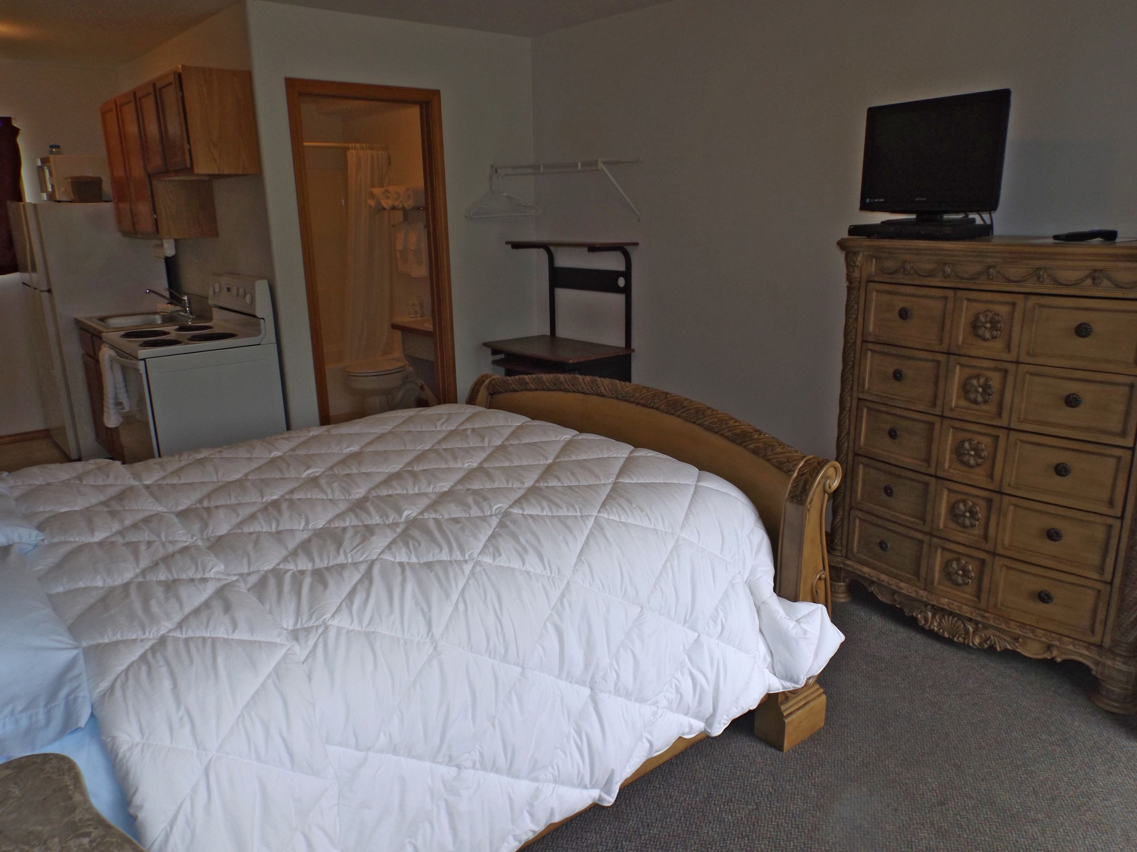 A bedroom with a large bed, a wooden dresser, a television, and a kitchenette.