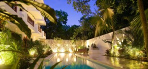 A pool with a stone wall and lush greenery, a wooden bench, and a white building with windows. - Livetulum (Tulum)