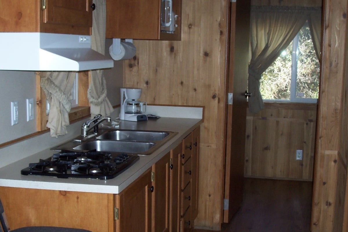 A compact kitchen with wooden cabinets, a gas stove, a sink, and a window with curtains.