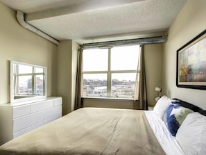 Un dormitorio con cama, cómoda, ventana con vistas a edificios y un cuadro en la pared.
