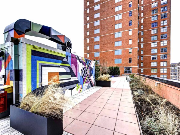 Terraza en la azotea con un mural colorido, plantas en macetas y vista de un edificio de ladrillo alto.