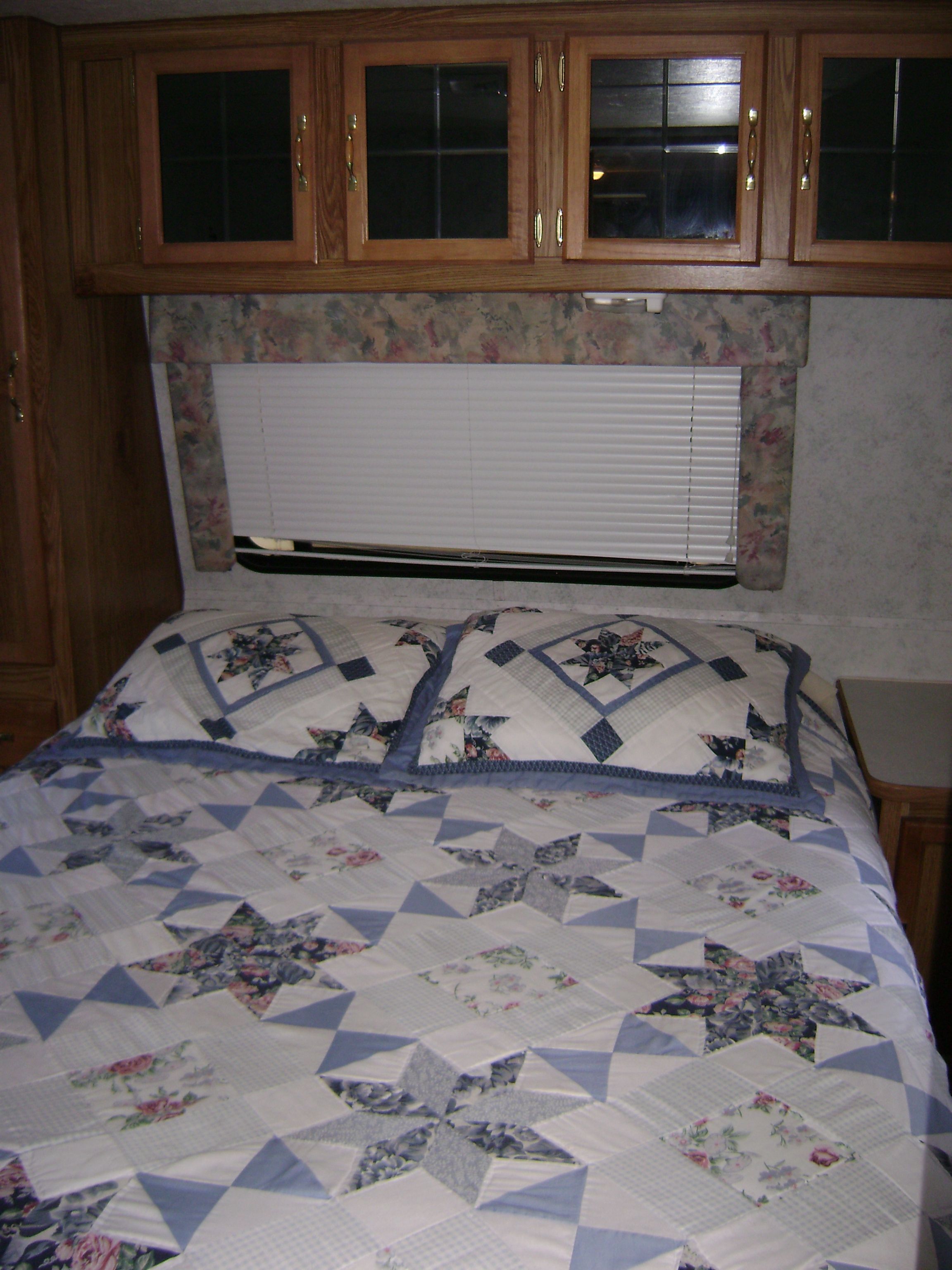 A bedroom with a bed, floral-patterned curtains, wooden cabinets, and a window with blinds.