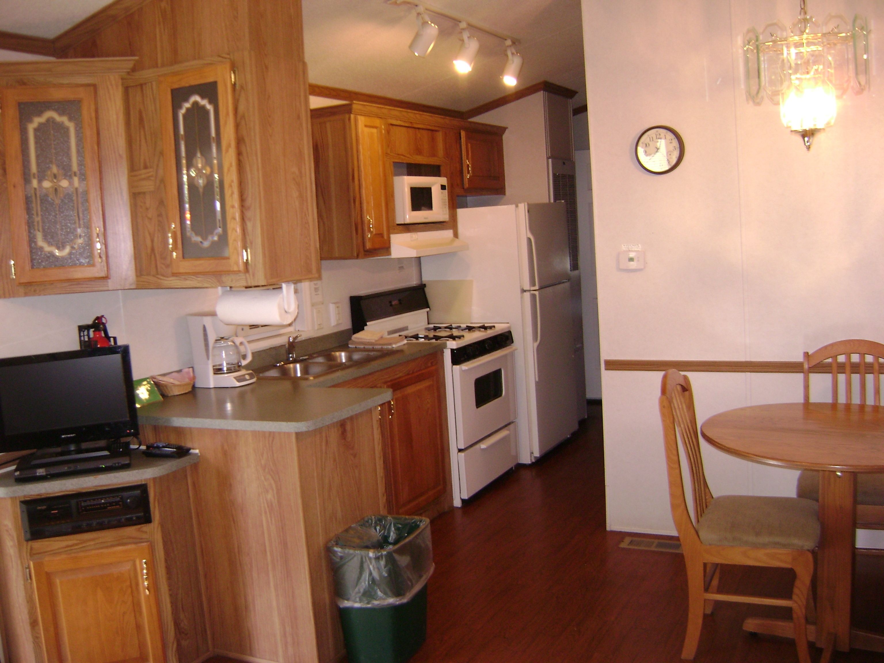 A kitchen with wooden cabinets, a microwave, a stove, a refrigerator, a trash bin, and a dining table with a chair.