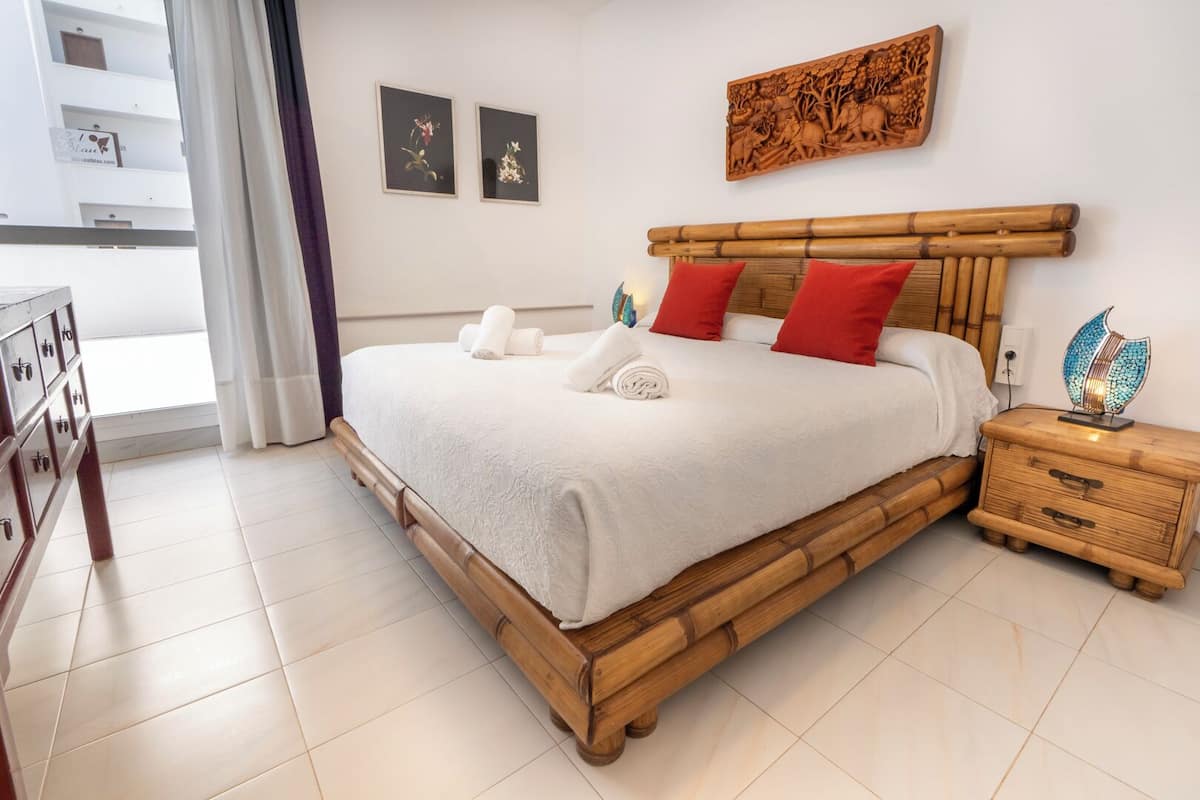 A neatly made bed with white linens and red pillows, a wooden nightstand with a decorative lamp, and two framed artworks on the wall.