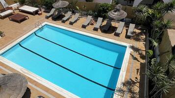 A rectangular swimming pool with two lanes, surrounded by lounge chairs and umbrellas, with a tiled deck and potted plants.