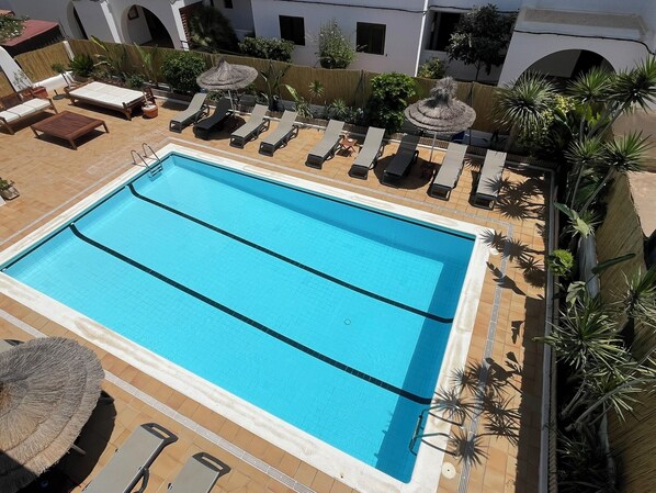 A rectangular swimming pool with two lanes, surrounded by lounge chairs and umbrellas, with a tiled deck and potted plants. - Apartamentos Cel Blau (Santa Eulalia del Rio)