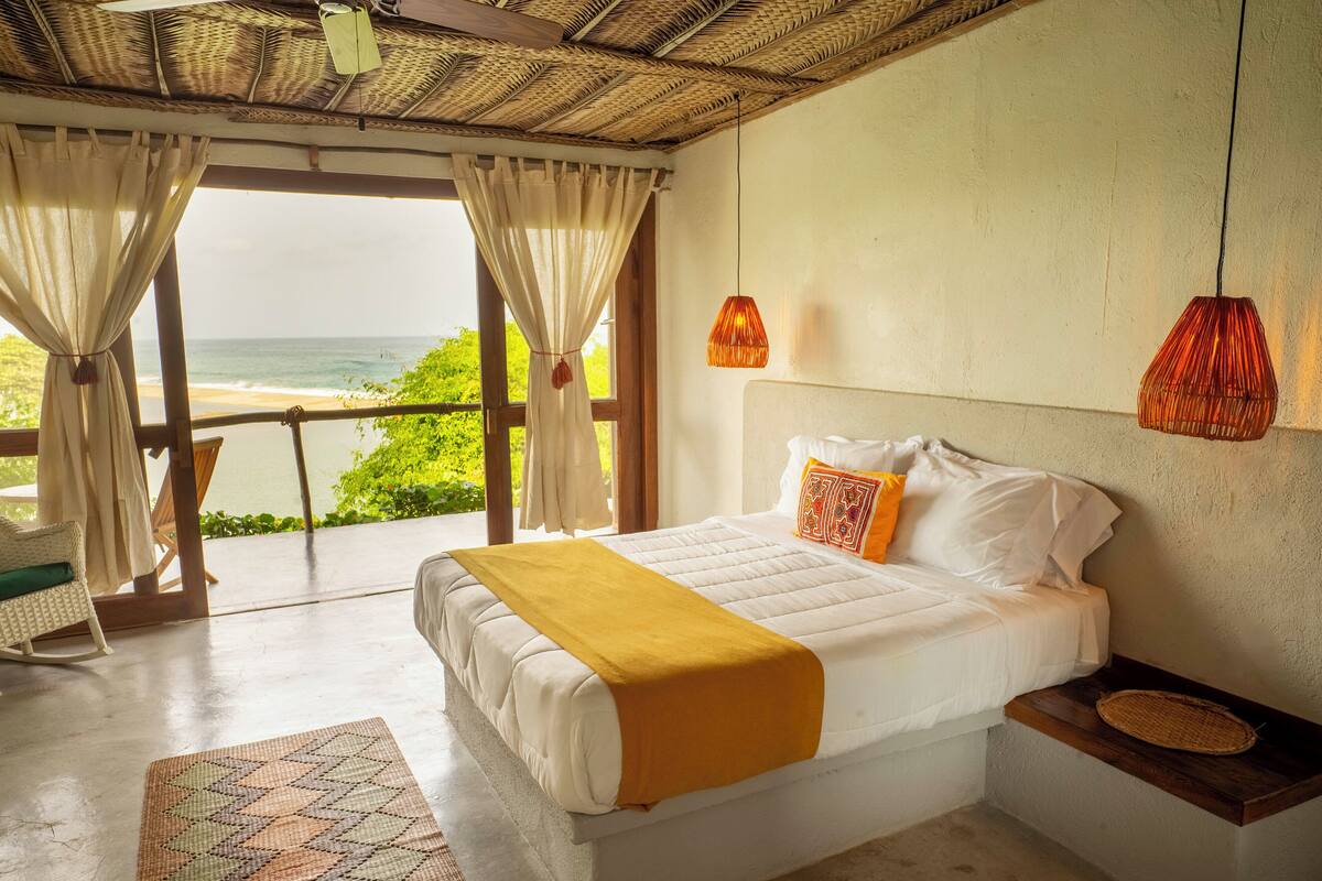 Un dormitorio con cama, mesita de noche, una cesta tejida y vistas al océano a través de amplias ventanas.