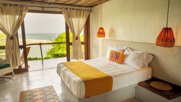 A bedroom with a bed, a nightstand, a woven basket, and a view of the ocean through large windows.
