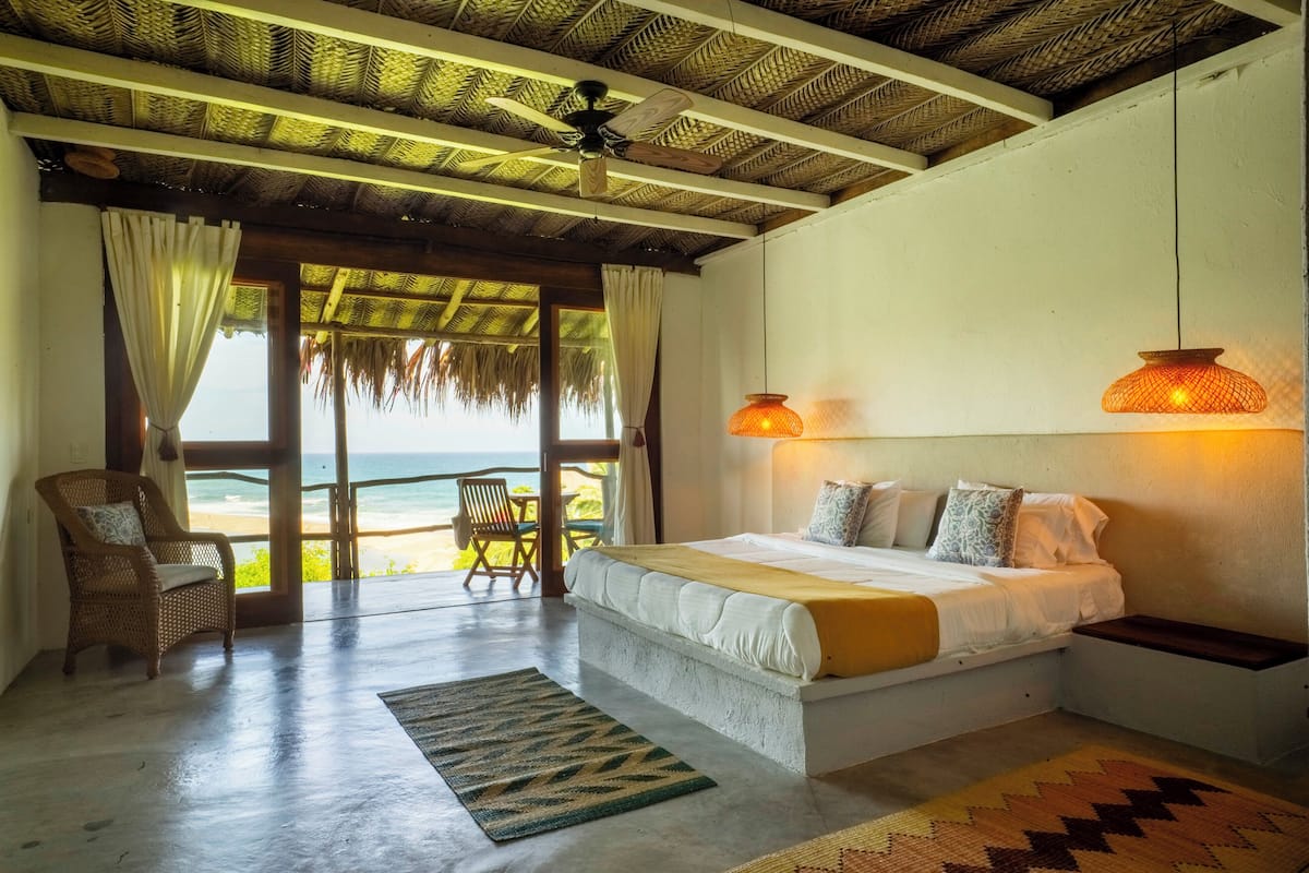 Un dormitorio con una cama grande, ventilador de techo, vista a la playa y un comedor con mesa y sillas.