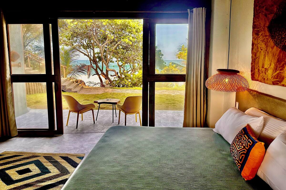 Habitación de hotel con cama, vista a la playa y zona de comedor con sillas y mesa.