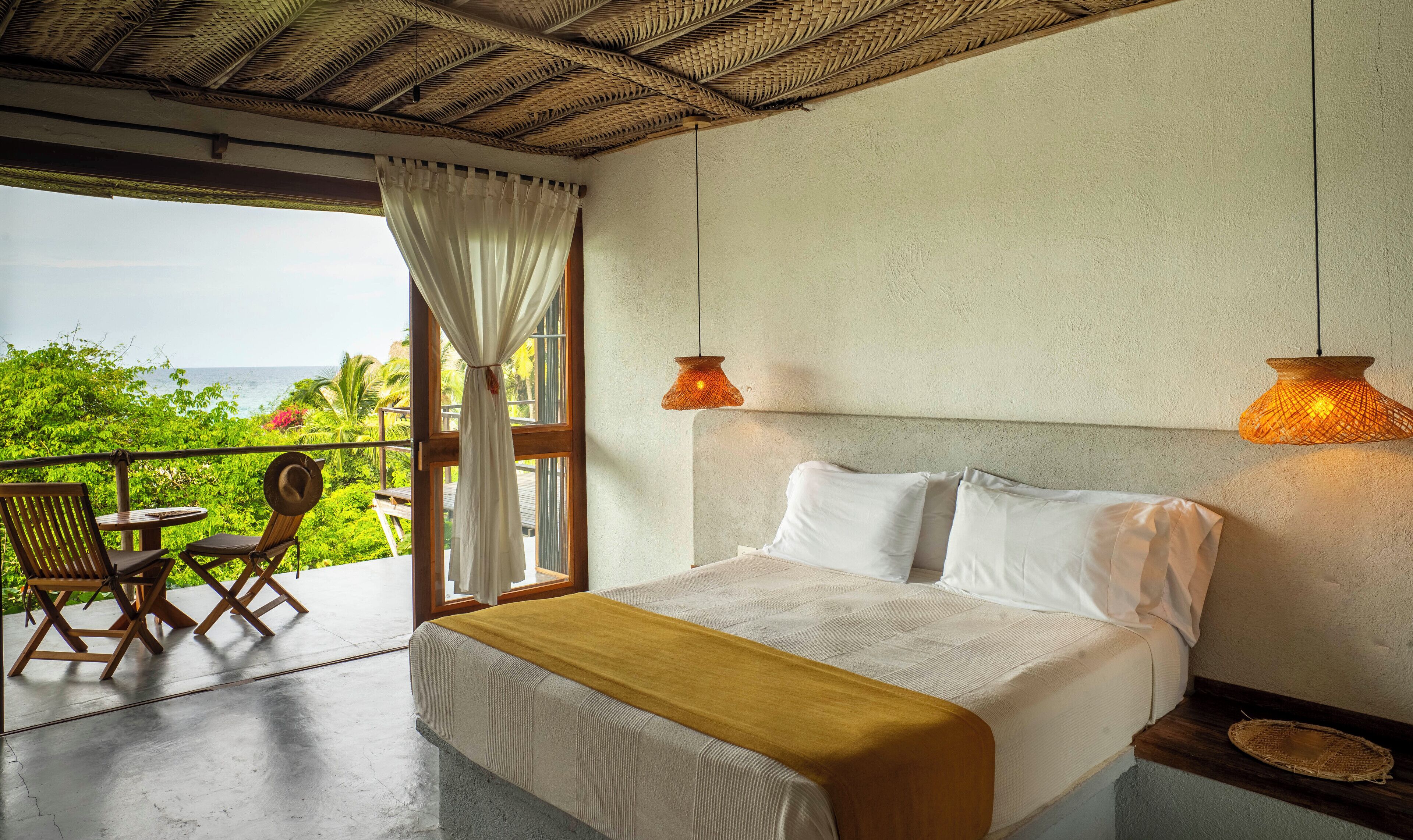 A bedroom with a bed, a view of the ocean, a balcony with a table and chairs, and two wall-mounted lamps.
