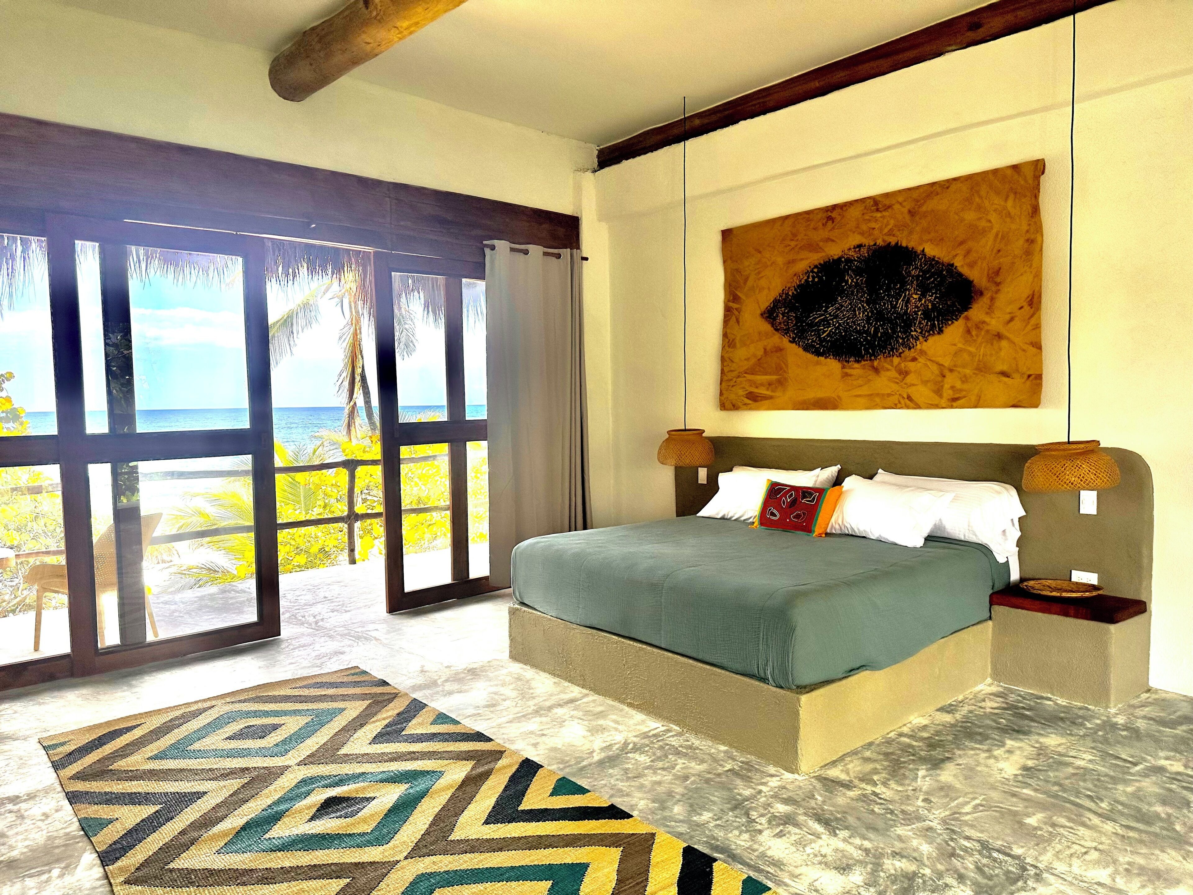 Un dormitorio con una cama grande, vista al océano a través de una puerta corredera y un cuadro con motivos tropicales en la pared.