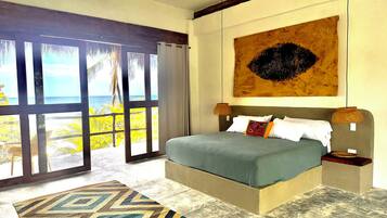 Un dormitorio con una cama grande, vista al océano a través de una puerta corredera y un cuadro con motivos tropicales en la pared.