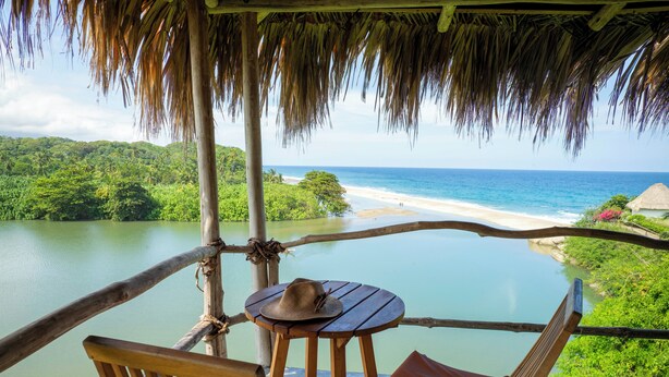 Una terraza de madera con dos sillas y una mesa, con vista a una playa y una zona boscosa.