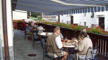 An outdoor patio area with tables and chairs, a covered seating area, and a view of a building with a white facade and red flowers.