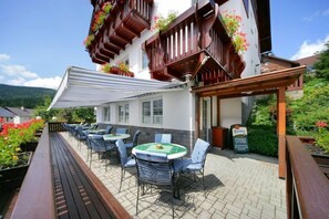 An outdoor patio with tables and chairs, a wooden deck, and a building with balconies adorned with flowers. - Pension Alba (Spindleruv Mlyn)