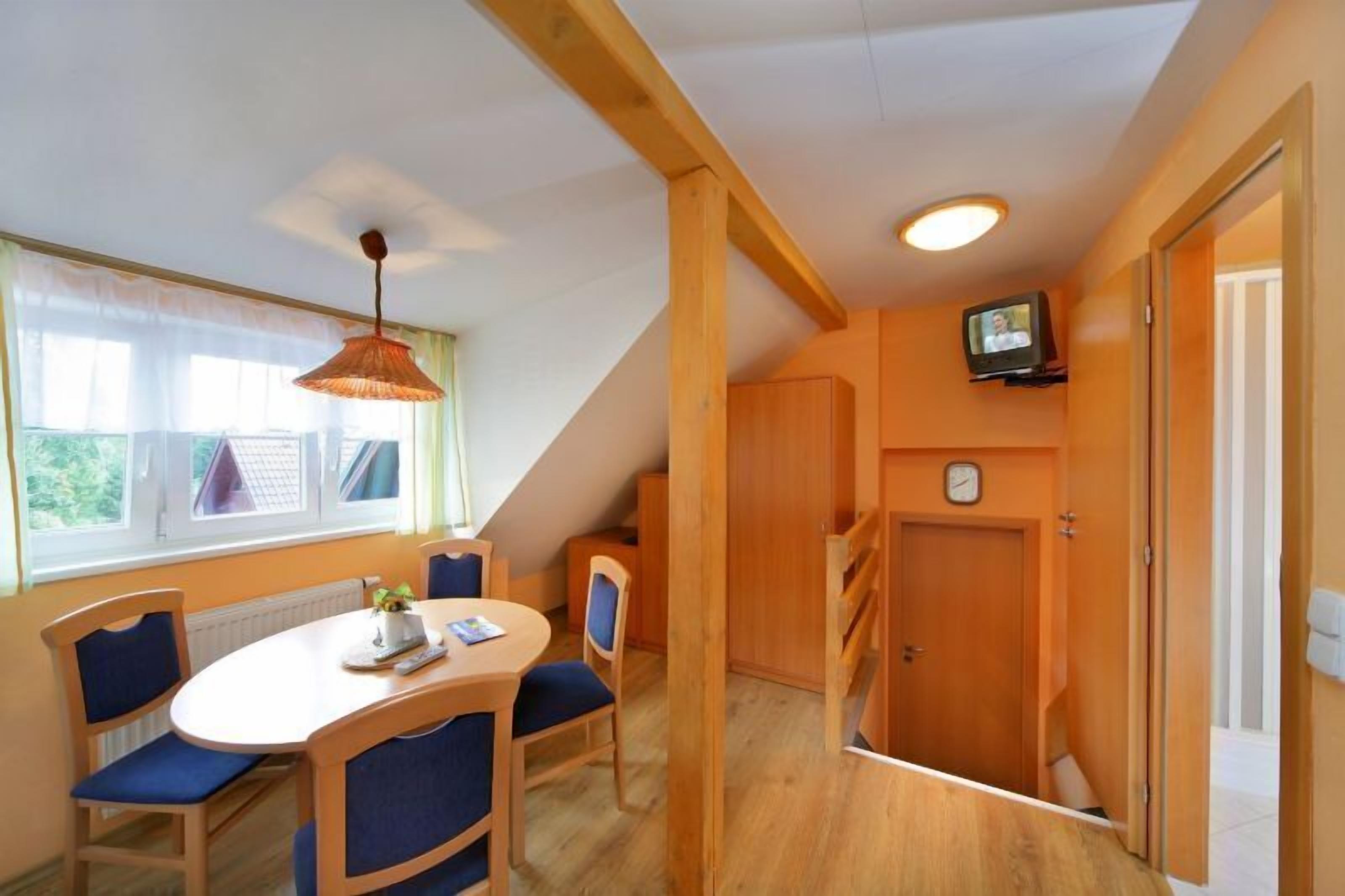 A cozy room with a wooden dining table, blue chairs, a hanging lamp, a television on the wall, and a clock.