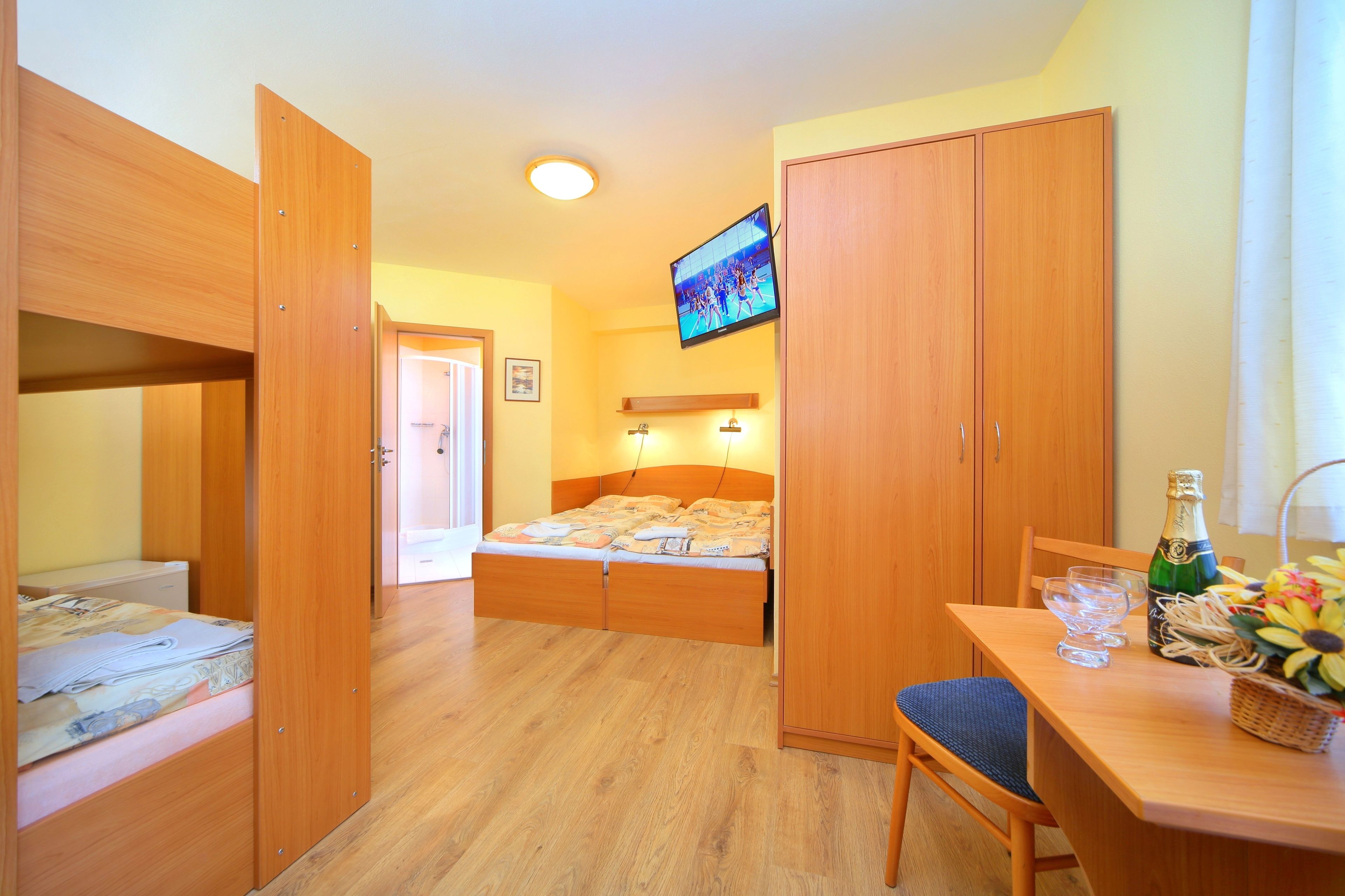 Een kleine kamer met een stapelbed, een bureau met stoel, een aan de muur bevestigde televisie en een houten garderobe.