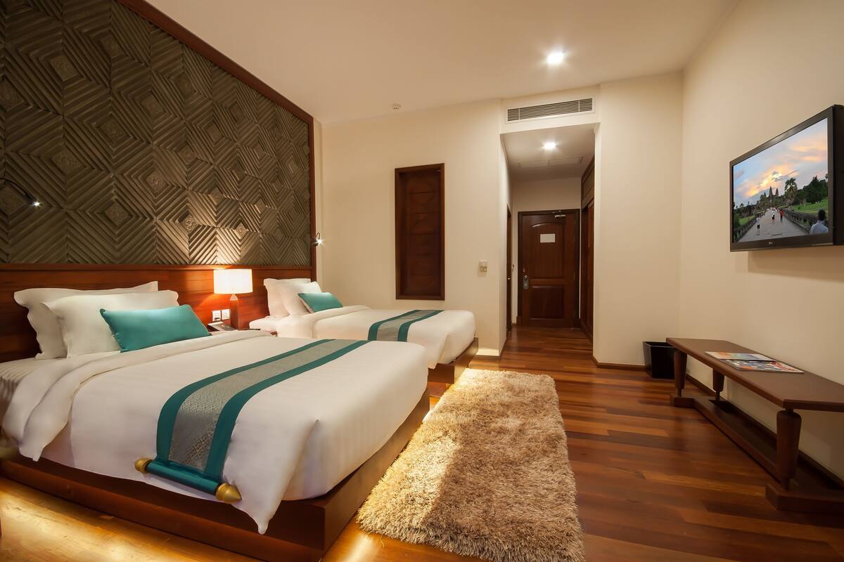 Une chambre d’hôtel avec deux lits, une tête de lit en bois, une grande fresque murale, un banc en bois et un tableau encadré au mur.