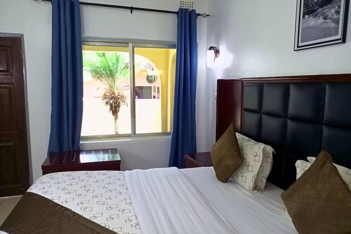 A hotel room with a bed, a nightstand, a window with curtains, and a wall-mounted picture.