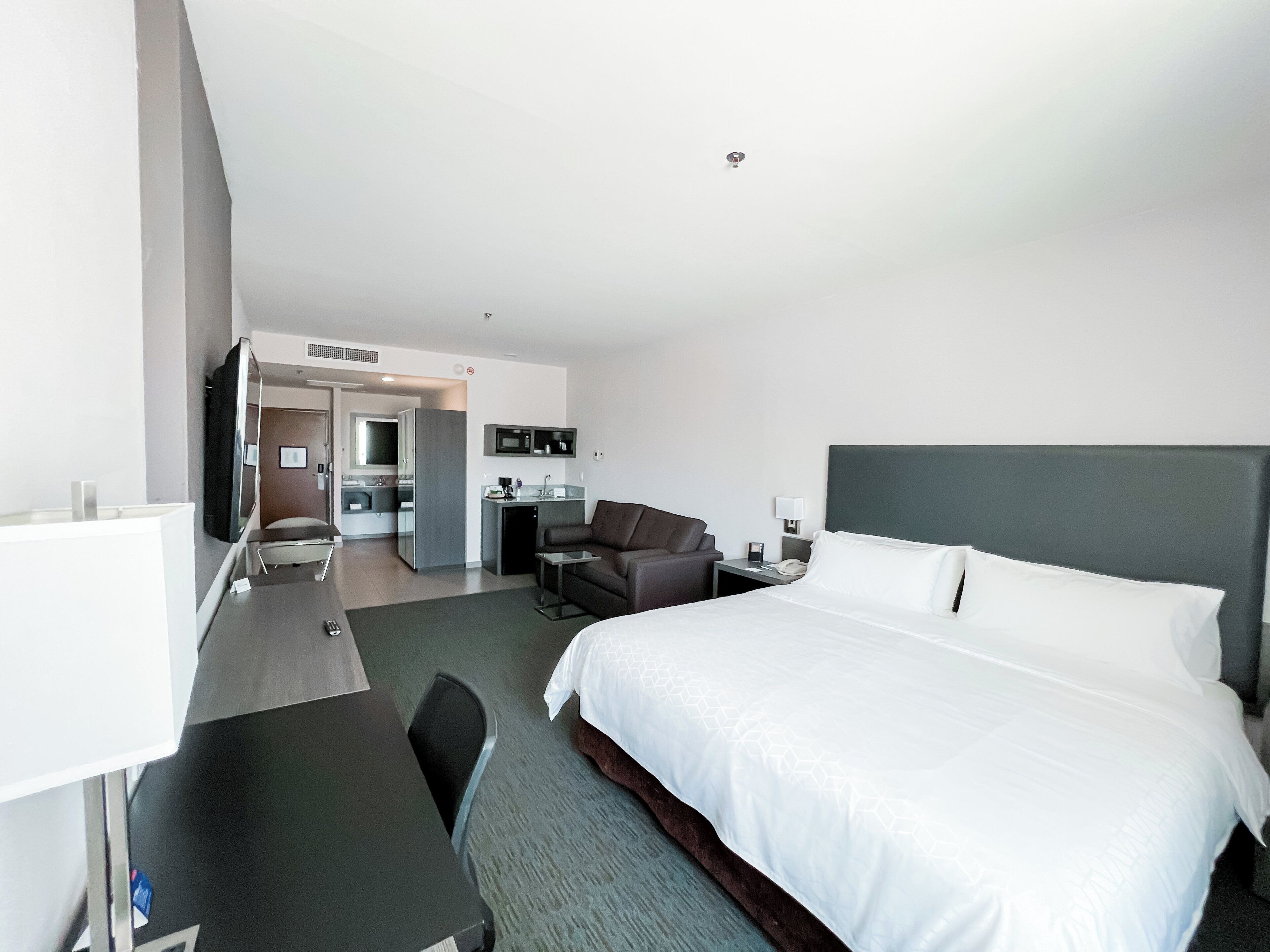 A modern hotel room with a large bed, a desk, a chair, a television, and a kitchenette.