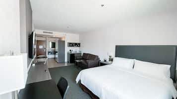 A modern hotel room with a large bed, a desk, a chair, a television, and a kitchenette.