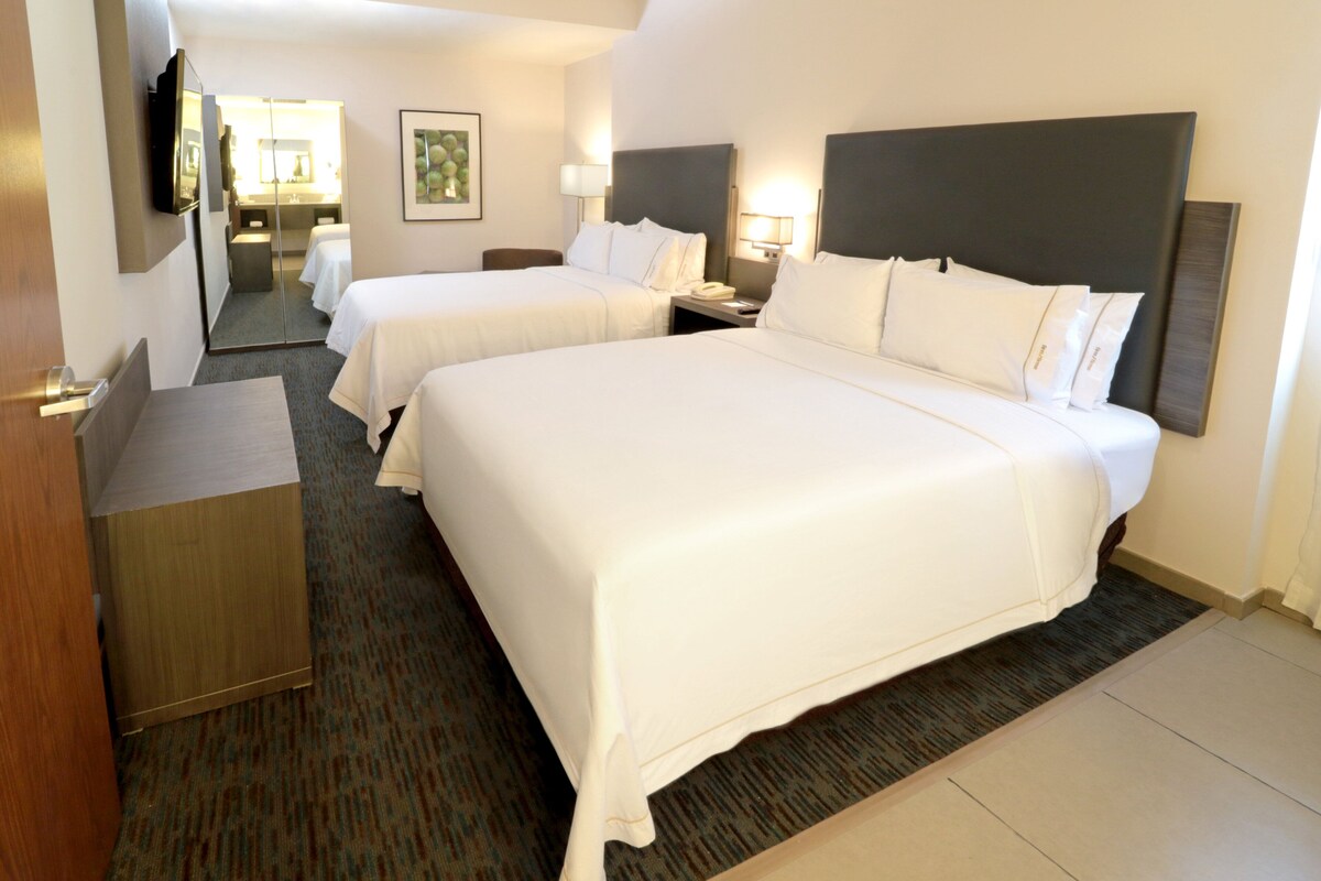 A hotel room with two beds, a desk, a mirror, and a nightstand with a lamp.