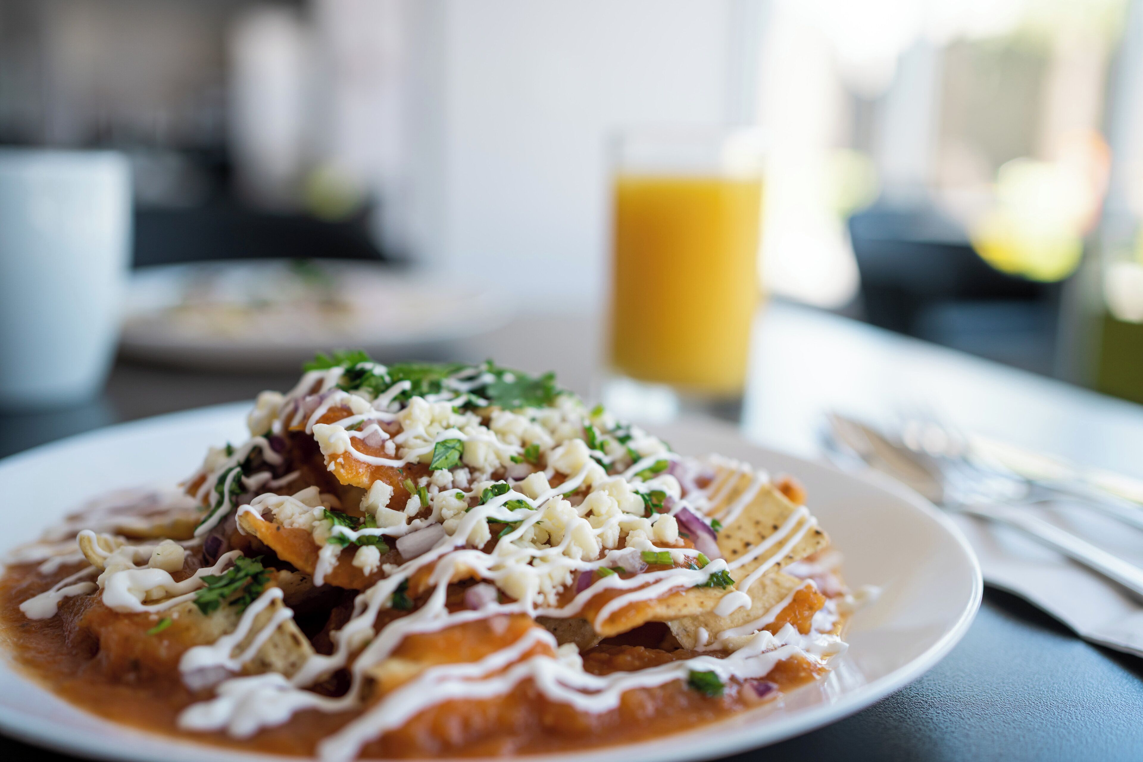 A plate of enchiladas topped with cheese, sour cream, and garnishes, with a glass of orange juice in the background.
