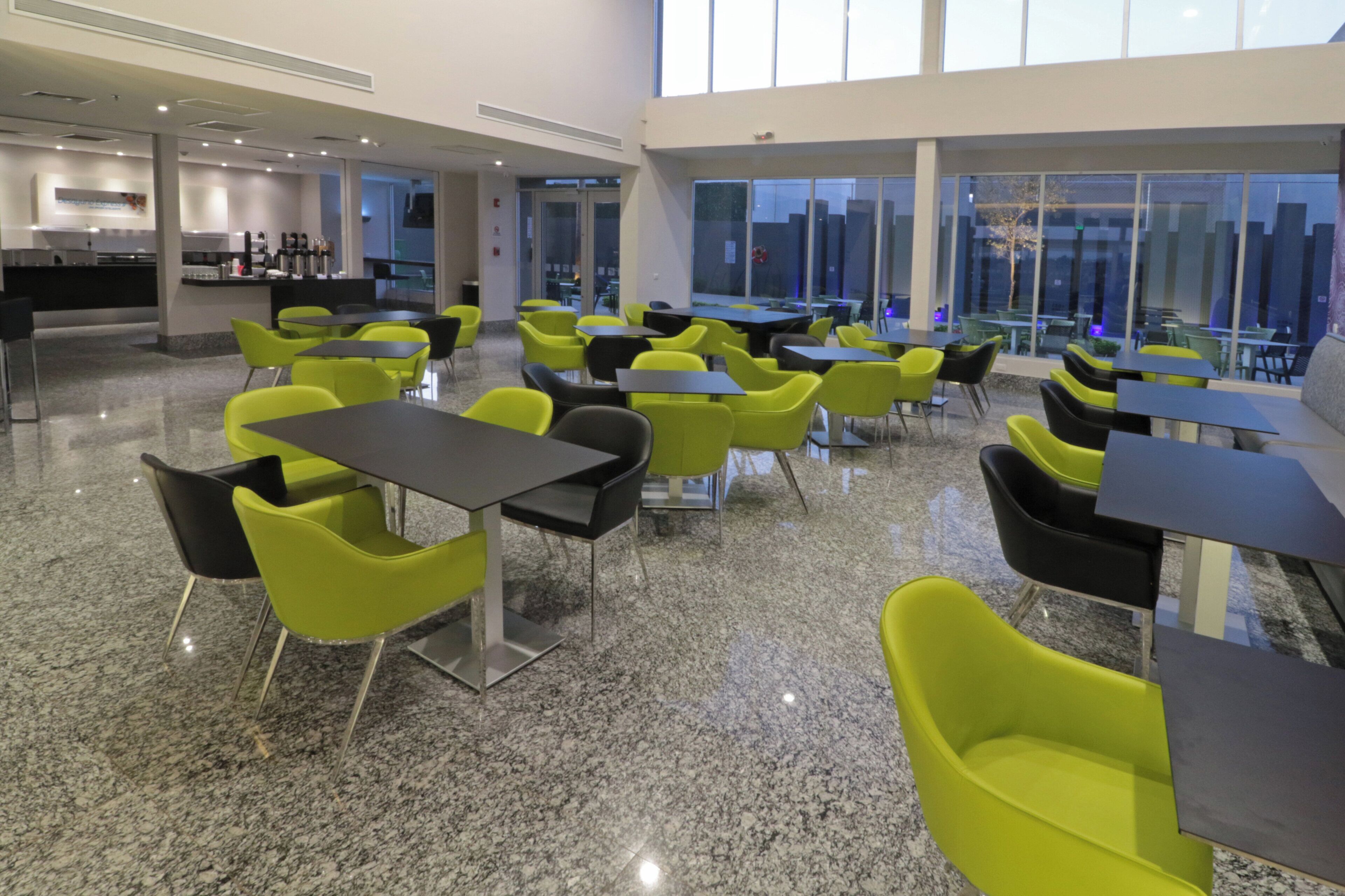 A modern indoor seating area with tables and chairs, a kitchen area in the background, and large windows providing a view of the cityscape.
