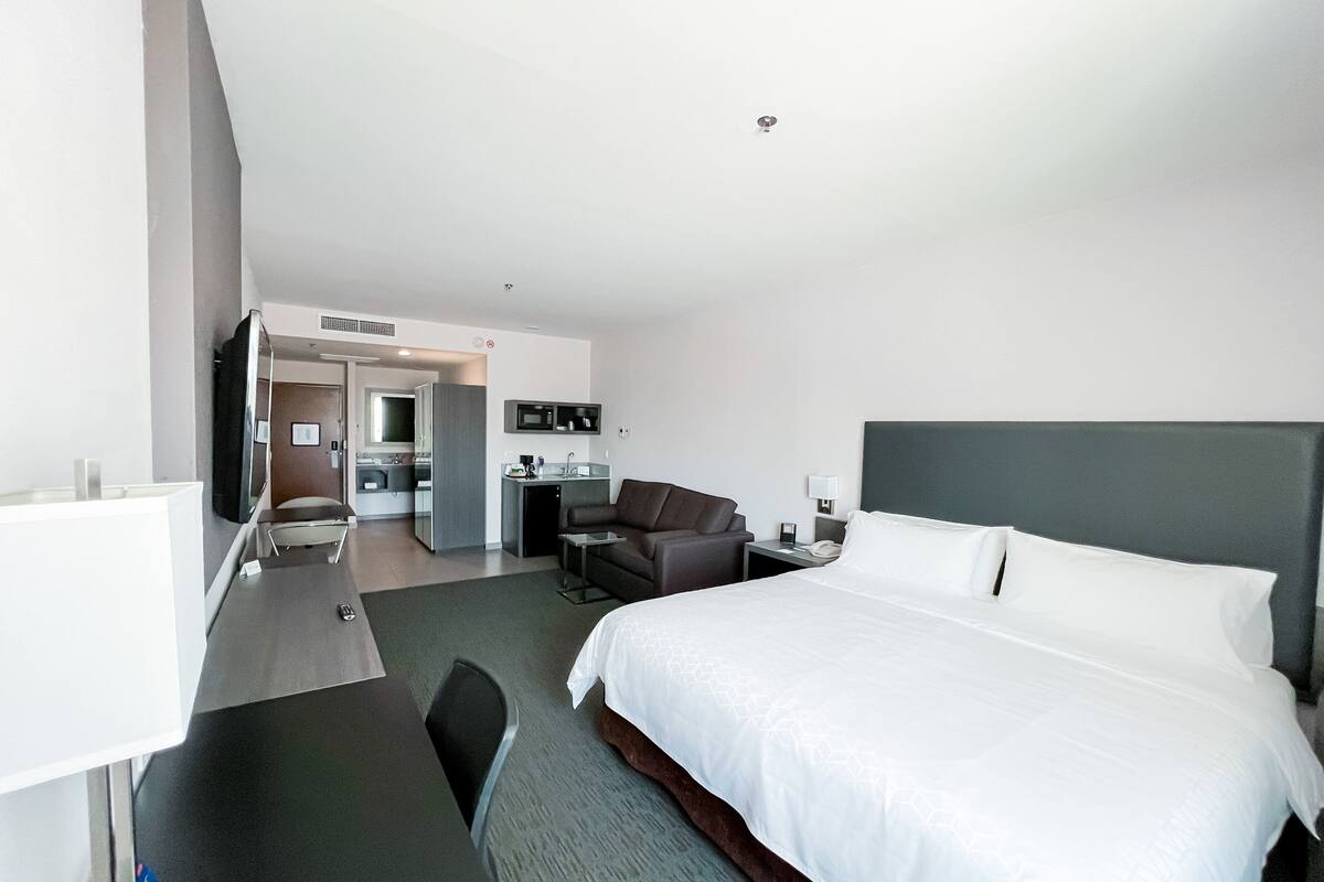 A modern hotel room with a large bed, a desk, a chair, a small kitchenette, and a TV.
