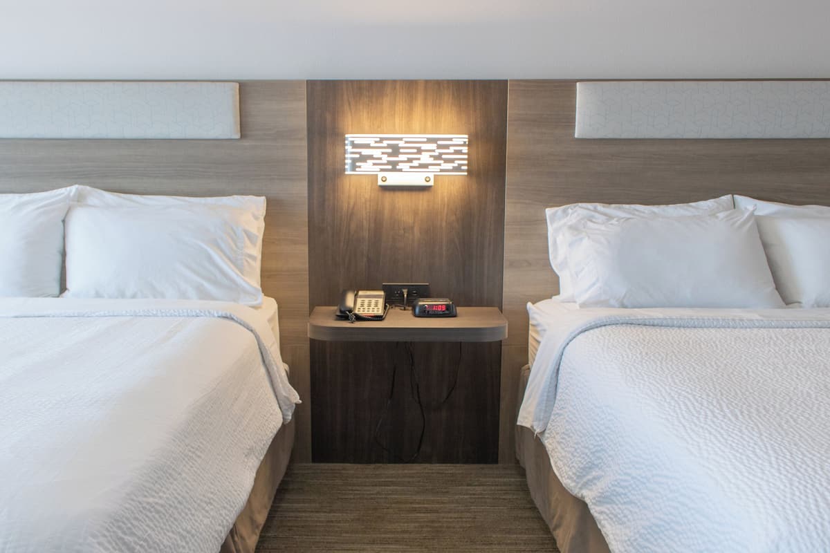 A hotel room with two beds, a bedside table with a phone and alarm clock, and a wall-mounted light fixture.