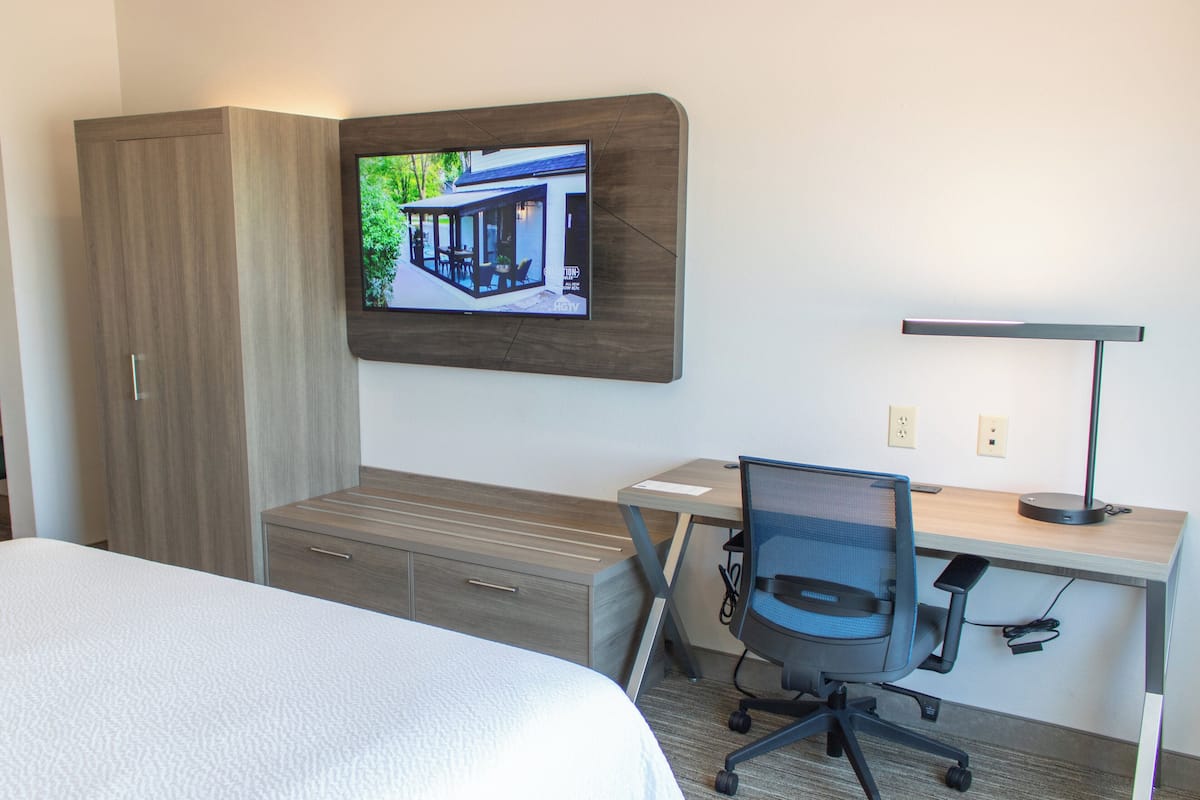 A hotel room with a bed, a desk with a chair, a TV mounted on the wall, and a wardrobe.