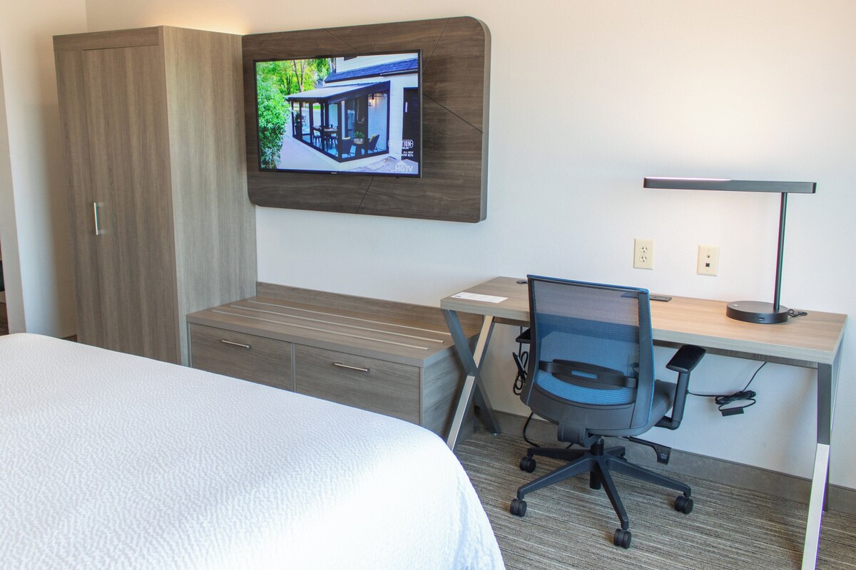 A hotel room with a bed, a desk with a chair, a TV mounted on the wall, and a wardrobe.