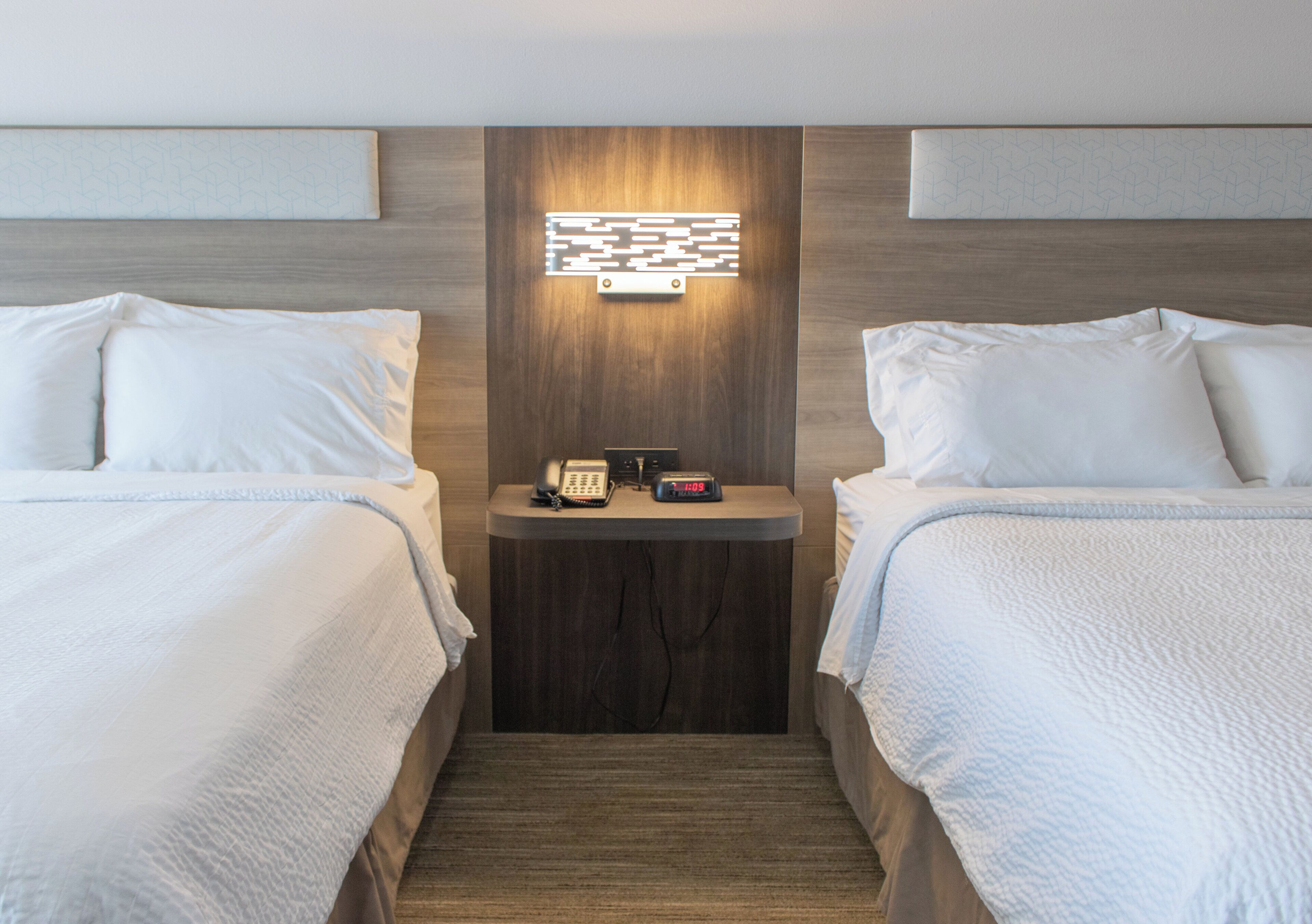 Habitación de hotel con dos camas, una mesita de noche con teléfono y despertador, y una lámpara de pared.