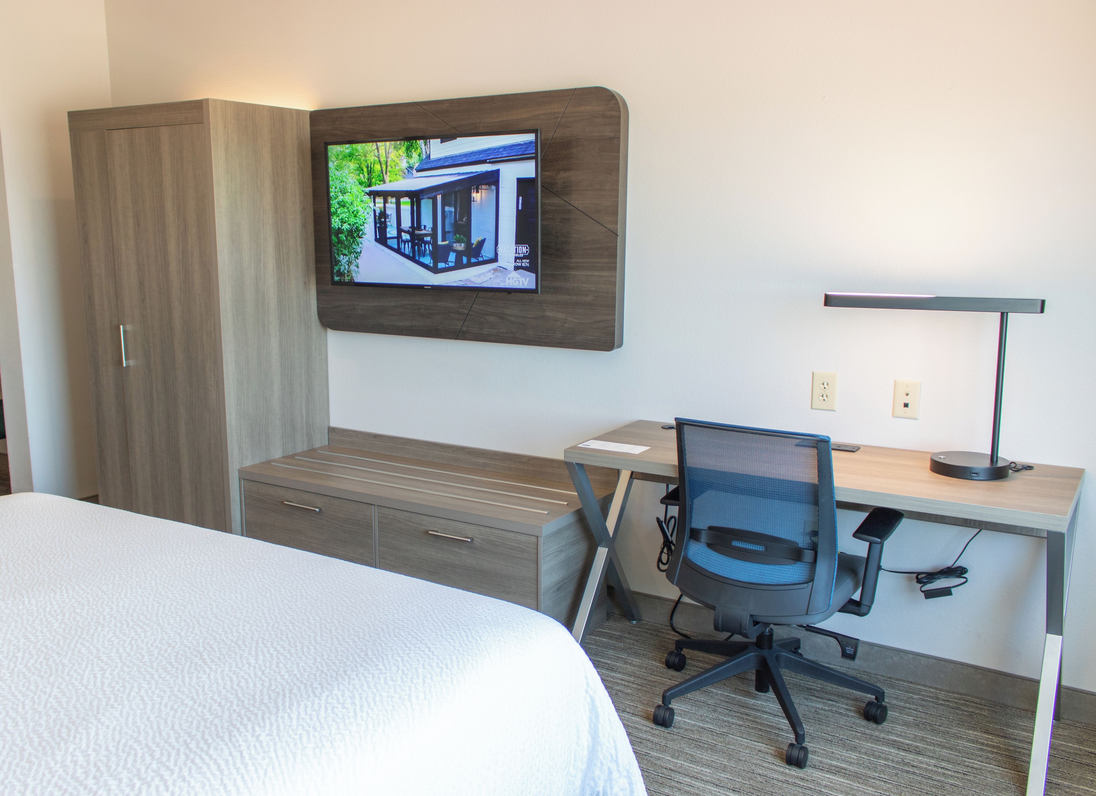A hotel room with a bed, a desk with a chair, a television mounted on the wall, and a desk lamp.