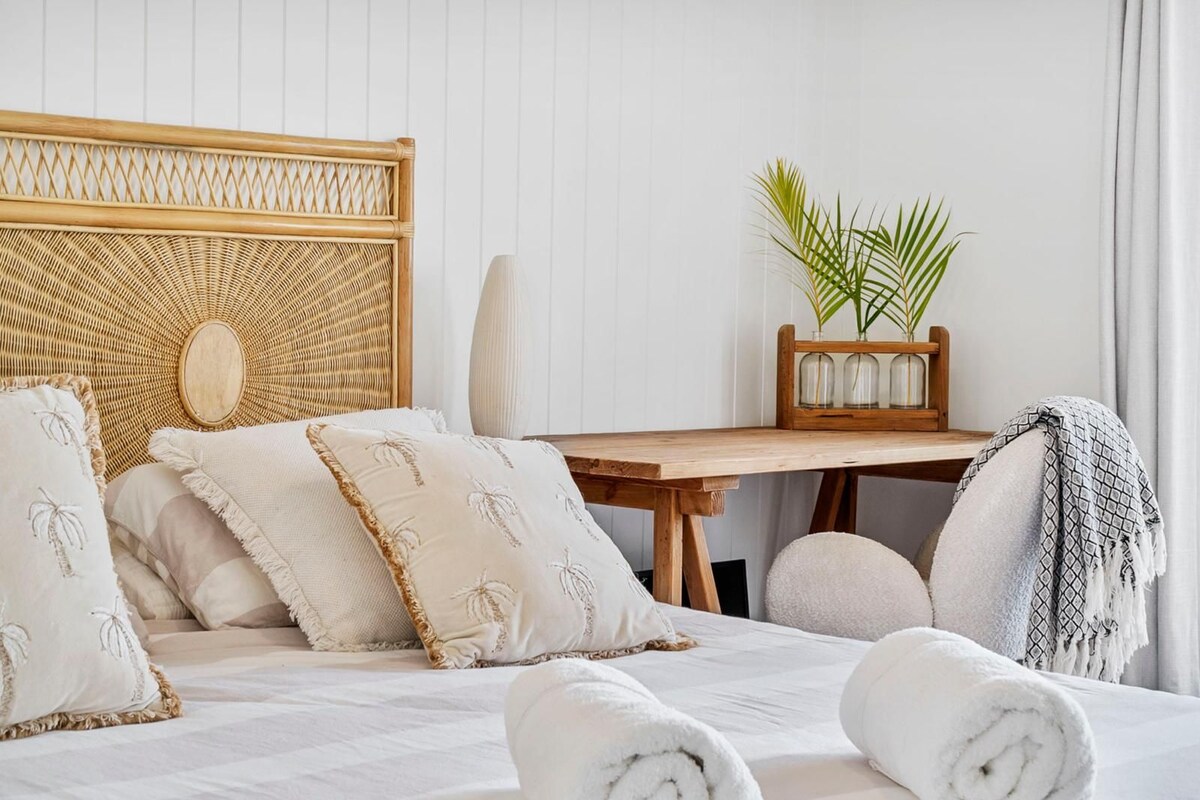Habitación de hotel con cama, almohadas, cabecera de madera, una mesa con planta y ventana con cortinas.