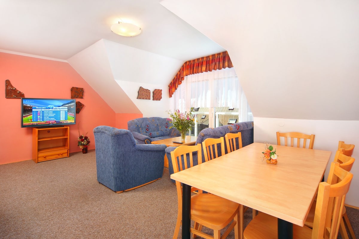 A cozy living room with a television, blue sofas, a wooden dining table, and a window with curtains.