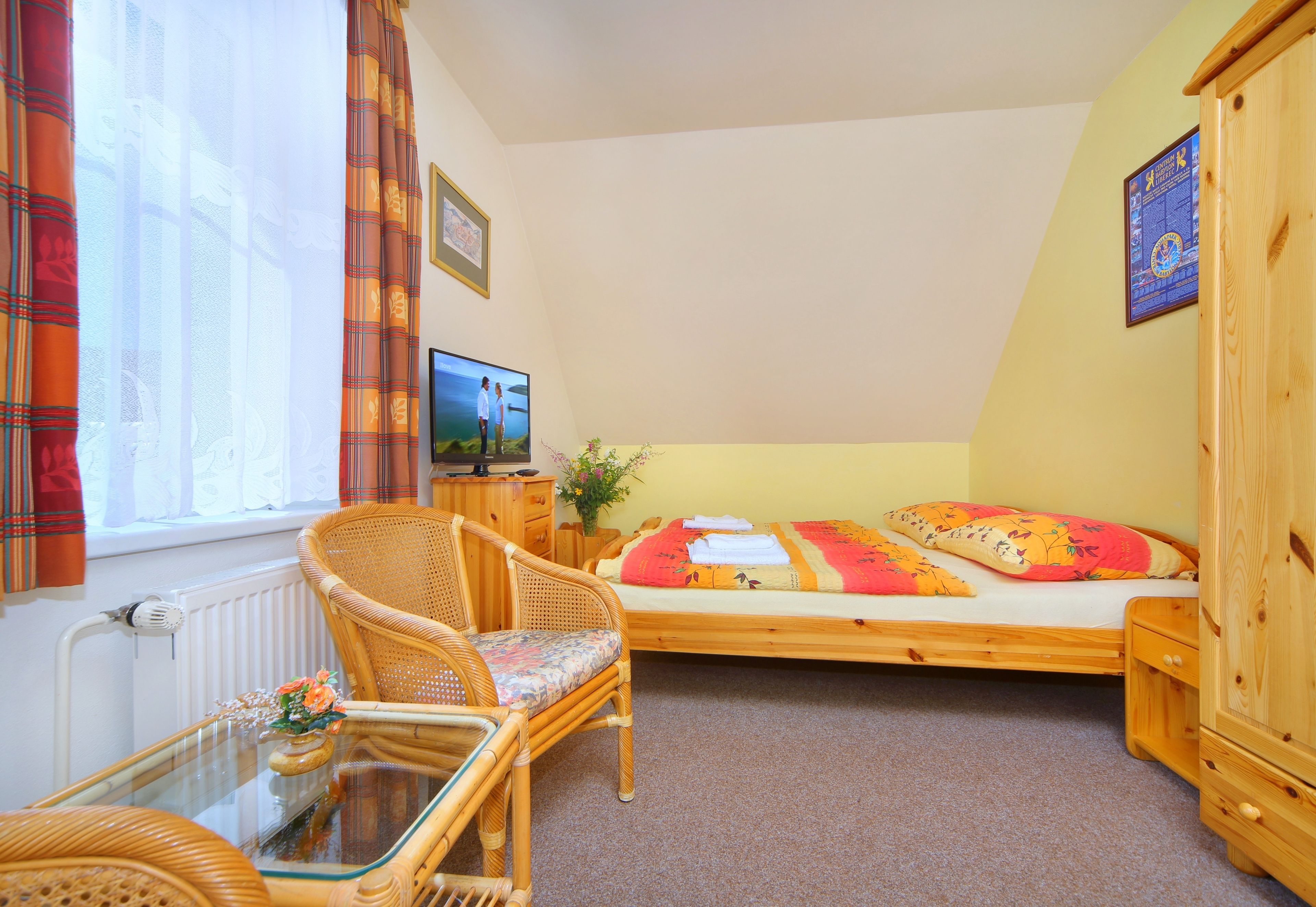 A small, cozy room with a single bed, a wicker chair, a glass-top table, and a television.