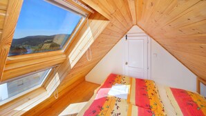 A small attic bedroom with wooden slats on the ceiling, a window with a view of a landscape, and a bed with a colorful blanket. - Pension Villa Stella (Spindleruv Mlyn)