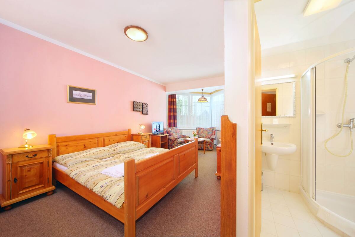 A bedroom with a wooden bed, bedside table with lamp, chair, and a view of the bathroom with a shower and sink.
