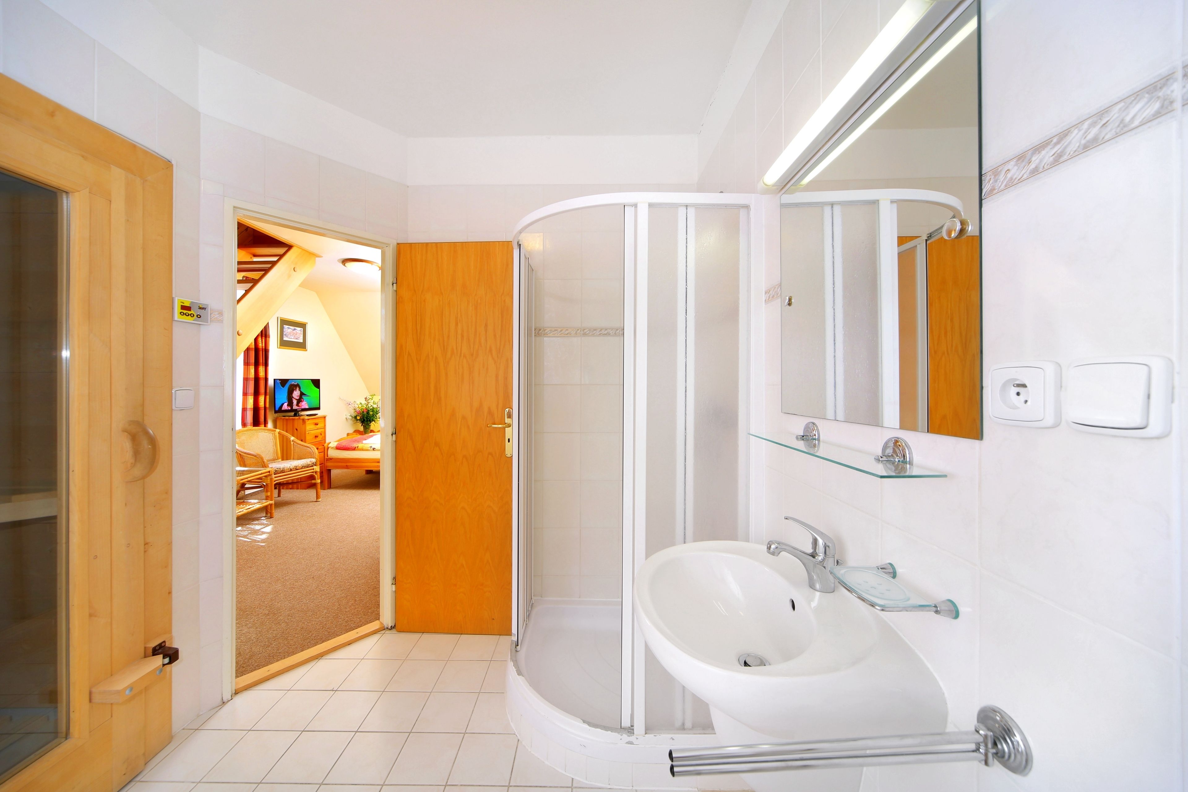 Ein modernes Badezimmer mit einer gläsernen Duschkabine, einem weißen Waschbecken und einer Holztür zu einem Wohnbereich.