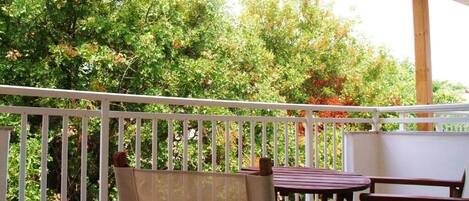 A balcony with a wooden table and chairs, surrounded by white railings, overlooking a lush green garden.