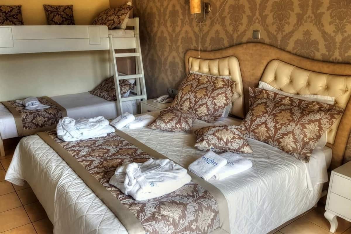 A hotel room with a bunk bed, a large bed with patterned bedding, and a bedside table with a lamp.