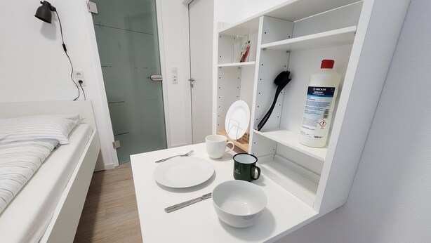 Una habitación pequeña y ordenada, con una cama, un escritorio blanco, un cuenco, una taza y una botella de líquido.