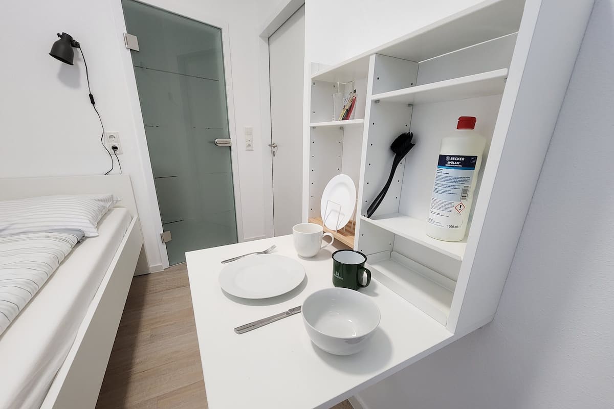 A small, neatly organized room with a bed, a white desk with a bowl, a mug, and a bottle of liquid.