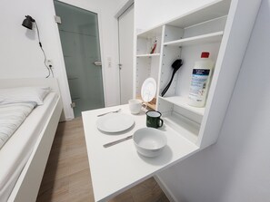 A small, neatly organized room with a bed, a white desk with a bowl, a mug, and a bottle of liquid.