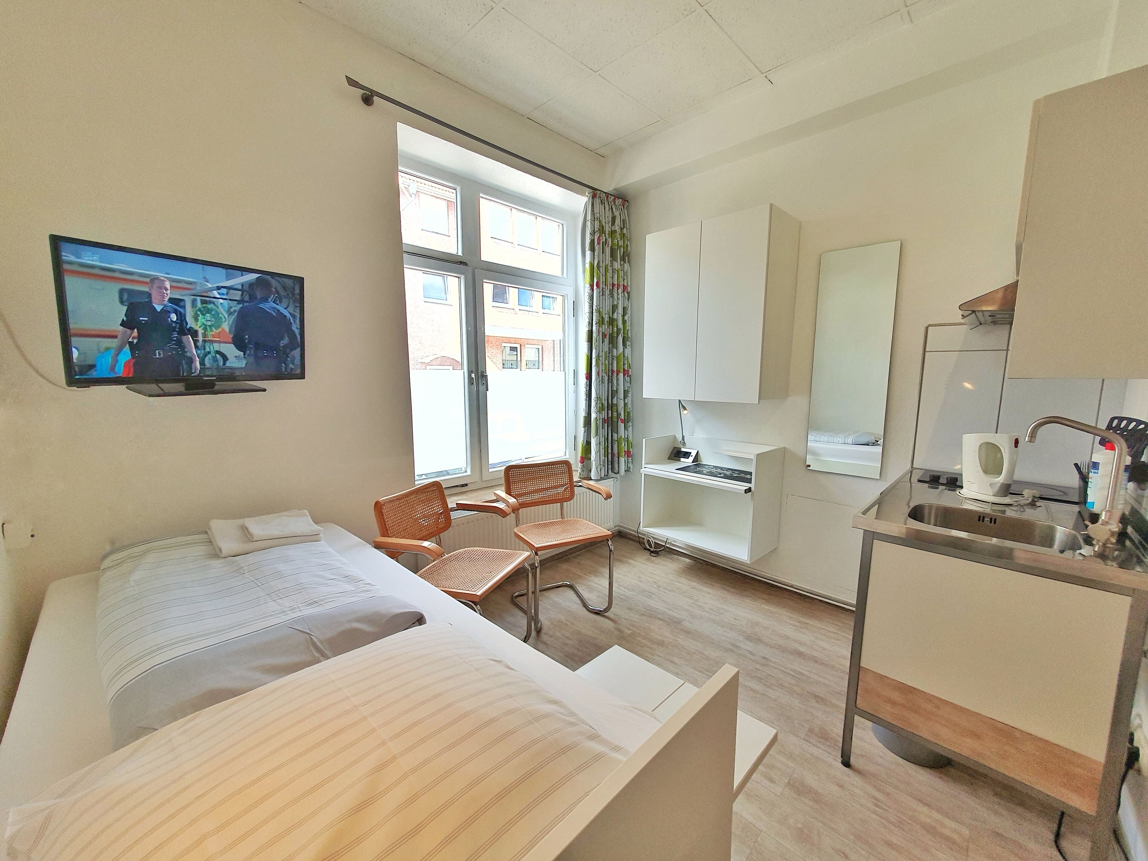A hotel room with a bed, a desk with a computer, a chair, a window with curtains, a television, and a sink.