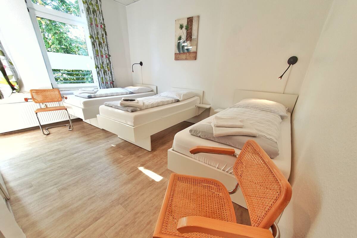 A modern bedroom with a large bed, a smaller sofa bed, a wooden chair, and a window with a view of greenery.