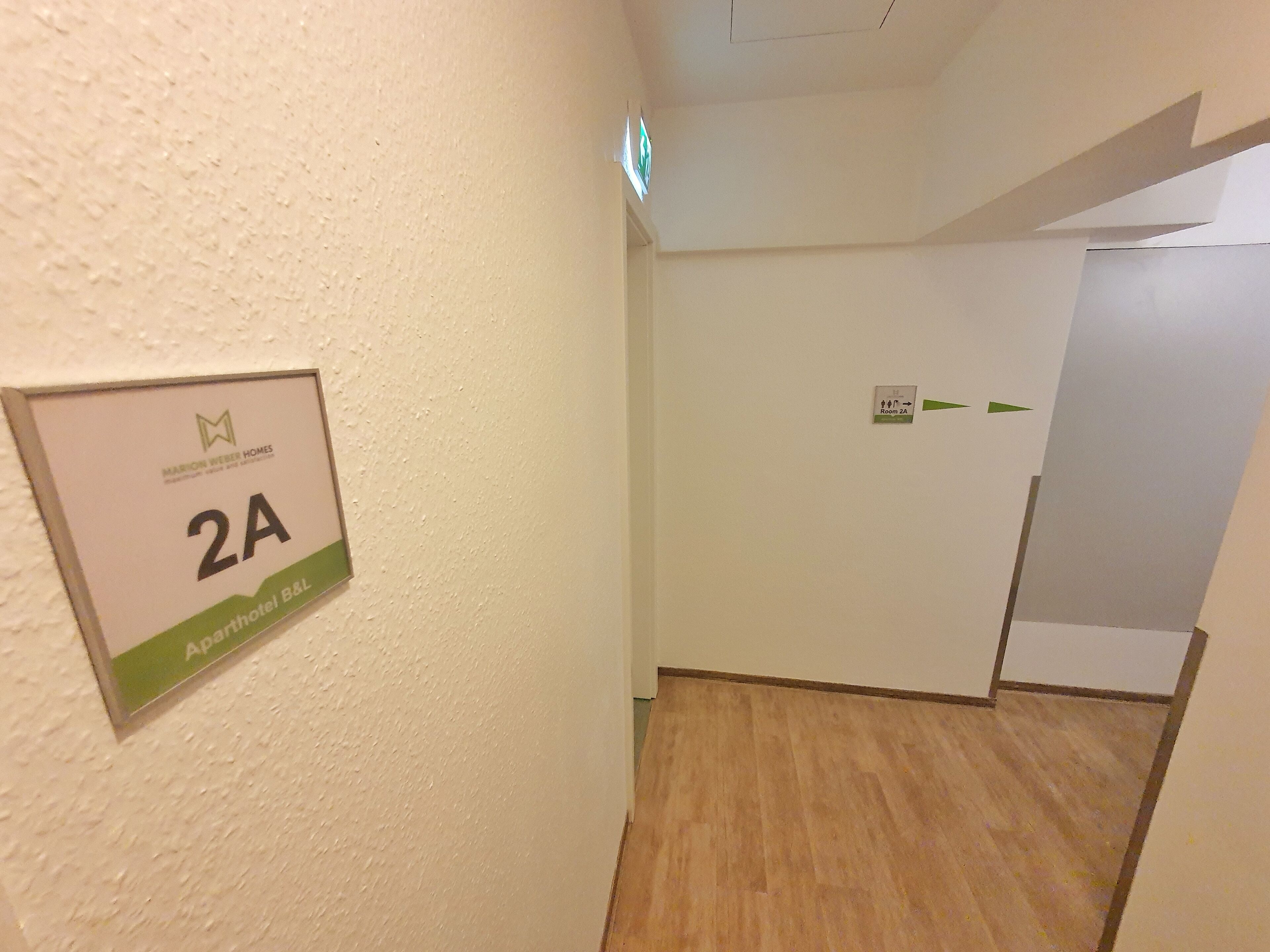 Ein Flur mit einem Schild, das 'Zimmer 2A' anzeigt, und Pfeilen, die zu 'Zimmer 2A' und 'Zimmer 2B' weisen.