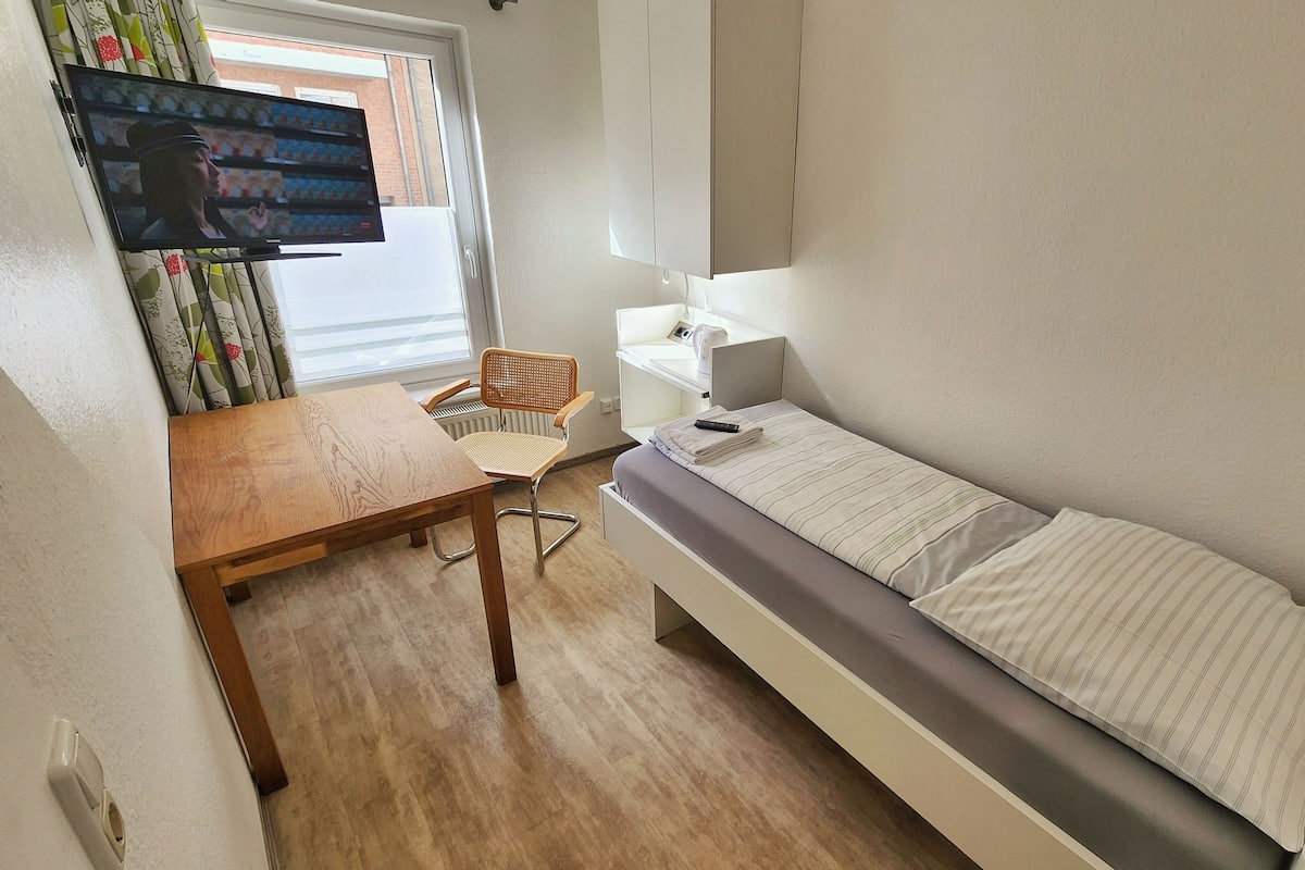 A small, neatly arranged room with a bed, a wooden table, a chair, a television, and a radiator.